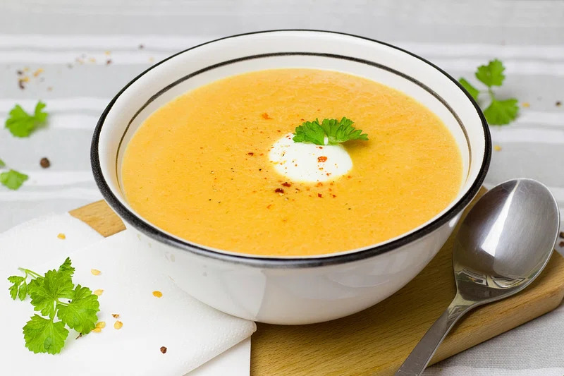 A hearty bowl of vegetable cauliflower soup featuring tender cauliflower florets, carrots, celery, and vibrant green herbs, served steaming hot in a white bowl with a slice of crusty bread on the side.