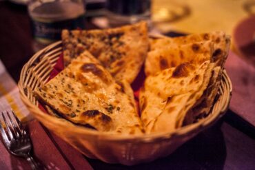 An assorted Indian breadbasket featuring a vibrant selection of naan, kulcha, missi roti, and tandoori paratha, served with bowls of flavorful curries, chutneys, and pickles on a rustic wooden platter, highlighting the rich diversity of Indian cuisine.