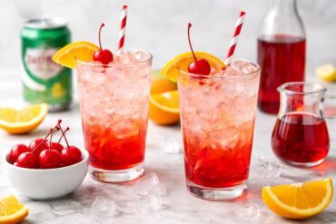 Two Shirley Temple mocktails in tall glasses with cherries and orange slices on top.