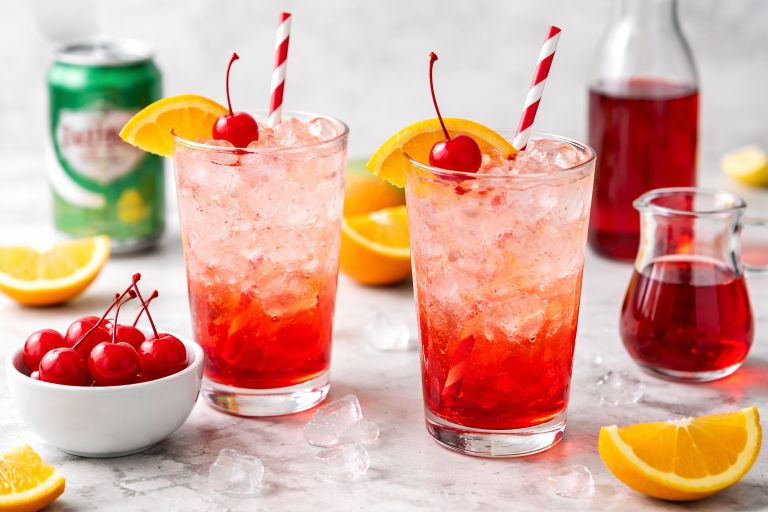 Two Shirley Temple mocktails in tall glasses with cherries and orange slices on top.
