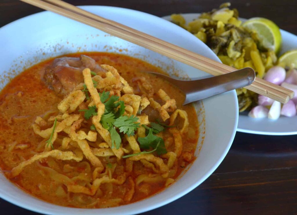 Traditional Thai Khao Soi: A bowl of creamy coconut curry noodle soup topped with crispy fried noodles, served with pickled mustard greens, red onions, and a wedge of lime on the side.
