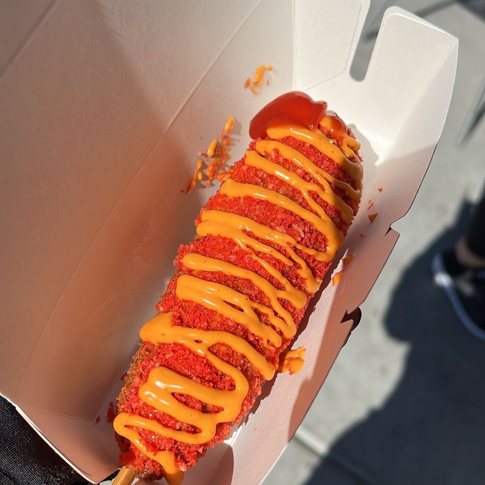 Close-up of Korean-style Veg corn dogs, featuring a golden, crispy batter with a light, fluffy texture. The corn dogs are filled with a savory vegetable mixture, coated in breadcrumbs, and drizzled with sweet and spicy sauces.