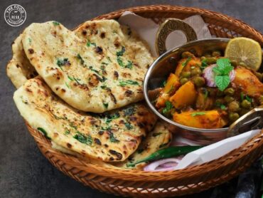 A freshly baked naan, golden and slightly charred, served on a wooden platter with butter melting on top, accompanied by a bowl of curry and garnished with cilantro.
