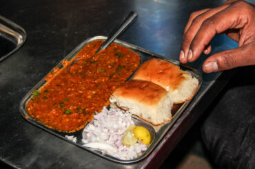 Plate of Pav Bhaji with a serving of spiced vegetable mash, garnished with chopped onions, cilantro, and a wedge of lime, served alongside buttered pav (bread rolls)