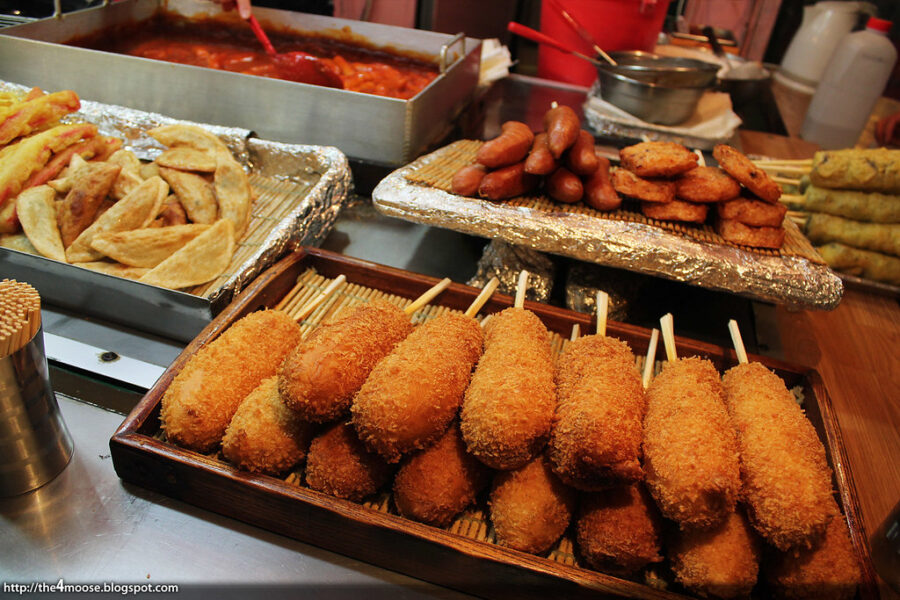 "Close-up of Korean corn dogs with an Indian twist, featuring crispy, golden-brown batter coated with a blend of Indian spices. The corn dogs are filled with a savory mix of vegetables, topped with tangy chutneys, and garnished with fresh cilantro and sesame seeds for a fusion of flavors.