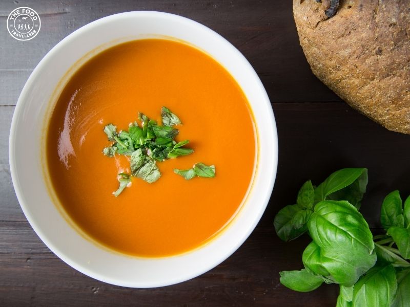 A bowl of creamy Roasted Tomato and Fennel Soup, garnished with a sprinkle of fresh herbs, showcasing a rich, smooth texture and vibrant orange-red color. The soup is served with a slice of toasted bread on the side.