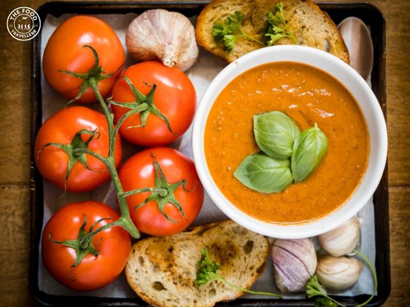 A bowl of Roasted Tomato and Fennel Soup, rich and smooth with a vibrant red color, served with whole garlic cloves and ripe tomatoes on the side. The soup is garnished with fresh herbs, and a slice of toasted bread accompanies the dish.