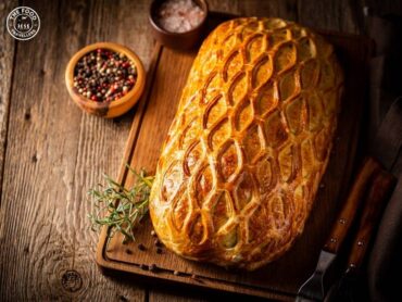 A golden, crispy Vegan Wellington with flaky puff pastry, sliced to reveal a hearty vegetable filling, served on a rustic wooden platter with fresh herbs for garnish.
