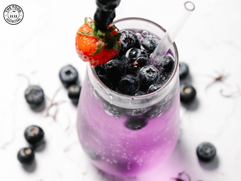 A refreshing Blueberry-Ginger Fizz Mocktail served in a tall glass, with ice cubes, fresh blueberries, a mint sprig, and a slice of ginger as garnish.