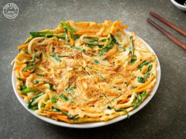 Close-up of a crispy Korean vegetable pancake, golden-brown and served with dipping sauce, garnished with fresh herbs.