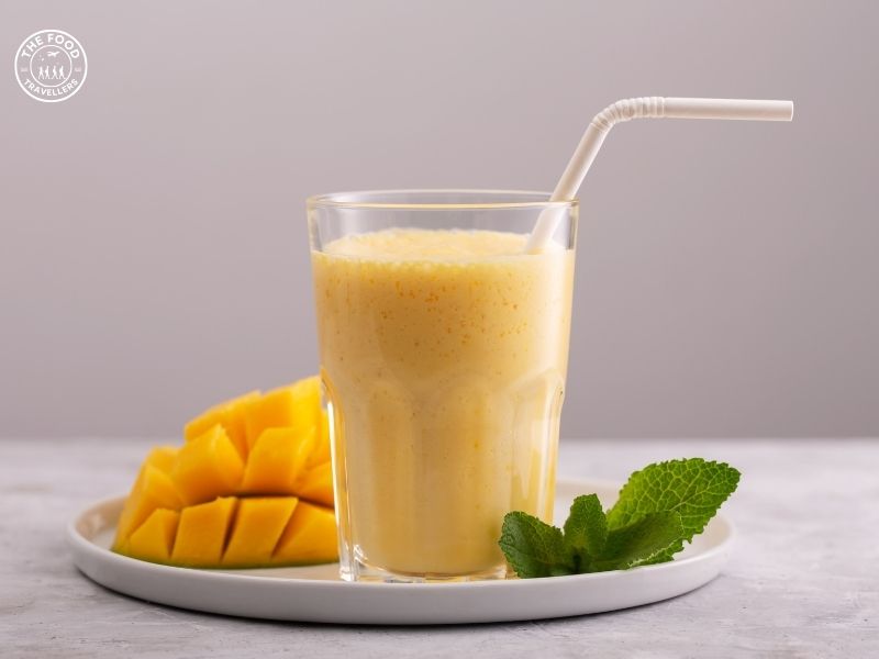 A glass of chilled mango lassi garnished with mint leaves and a sprinkle of cardamom, served on a wooden table.