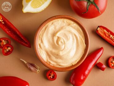 Creamy homemade chipotle sauce in a bowl surrounded by red chilies, lemon, and garlic on a neutral background