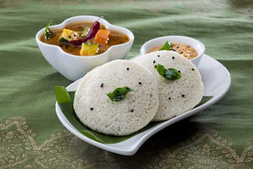 Idli recipe variations featuring steamed idli, thatte idli, podi idli, sambar idli, and mini button idli on rustic table